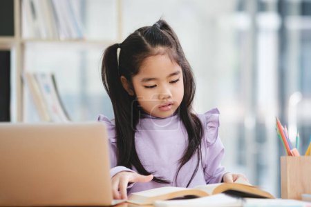 Foto de Una joven está sentada en un escritorio con un portátil y un libro. Ella está enfocada en su trabajo y está estudiando - Imagen libre de derechos