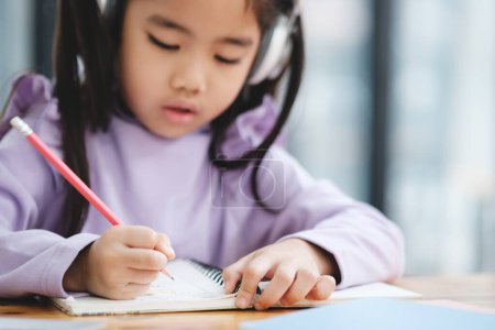 Téléchargez les photos : Une jeune fille écrit dans un cahier avec un crayon. Elle porte des écouteurs et se concentre sur son travail. La scène suggère qu'elle est engagée dans une tâche qui nécessite de la concentration - en image libre de droit
