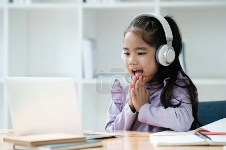Photo for A young girl wearing headphones is sitting at a desk with a laptop in front of her. She is smiling and she is enjoying herself - Royalty Free Image