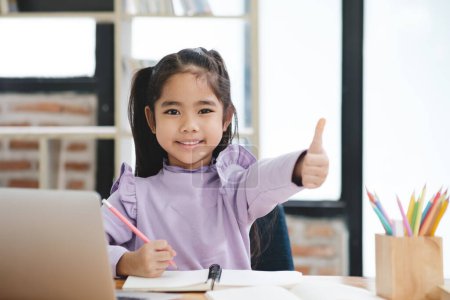 Photo for A young girl is sitting at a desk with a laptop and a notebook. She is smiling and giving a thumbs up. The scene suggests that she is happy and enjoying her work or activity - Royalty Free Image