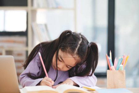 Photo for A young girl is sitting at a desk with a pencil and a book. She is writing in the book - Royalty Free Image