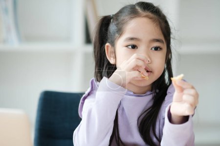 Photo for A young girl is eating a snack while sitting in a chair. She is wearing a purple shirt and has her hair in pigtails - Royalty Free Image