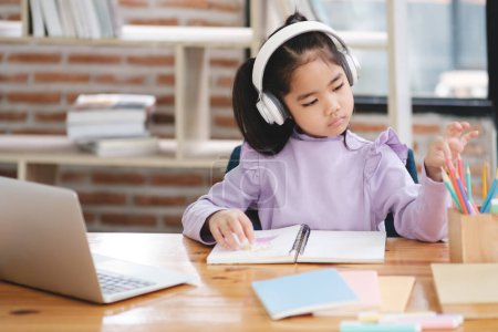 Téléchargez les photos : Une jeune fille est assise à un bureau avec un ordinateur portable et un cahier. Elle porte des écouteurs et se concentre sur son travail - en image libre de droit