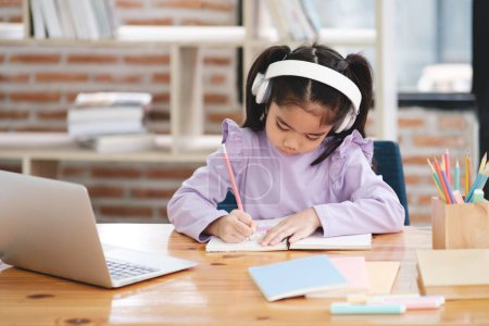 Photo for A young girl is sitting at a desk with a laptop and a notebook. She is writing in her notebook and wearing headphones. The scene suggests that she is working on a project or studying for an exam - Royalty Free Image