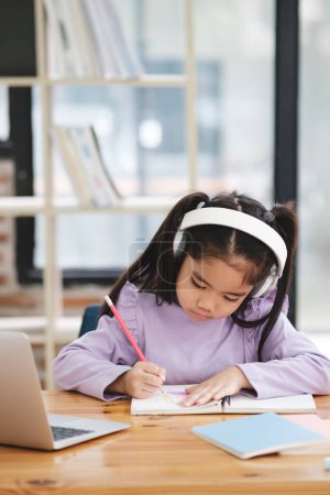 Photo for A young girl is sitting at a desk with a laptop and a notebook. She is wearing headphones and writing in her notebook - Royalty Free Image
