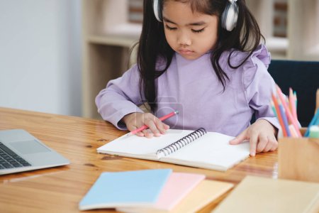 Photo for A young girl is sitting at a desk with a notebook and a laptop. She is wearing headphones and she is focused on her work - Royalty Free Image