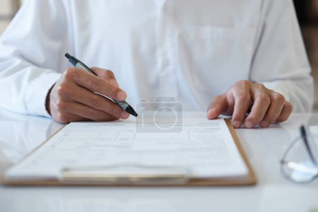 Photo for A man is writing on a piece of paper with a pen. He is wearing a white shirt and he is focused on his writing. Concept of concentration and determination as the man puts pen to paper - Royalty Free Image