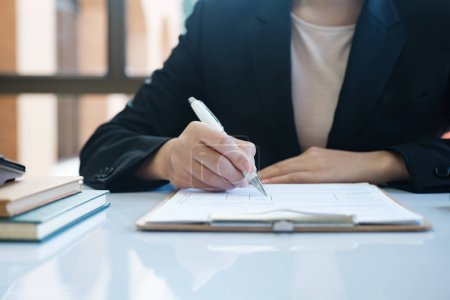 Photo for A woman in a suit is writing with a pen on a piece of paper. Concept of professionalism and focus, as the woman is likely working on a task or document - Royalty Free Image