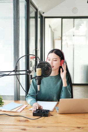 Photo for A woman is sitting at a desk with a microphone and a laptop. She is wearing headphones and she is recording a podcast or a voiceover. The room is well-lit and has a modern, professional feel - Royalty Free Image