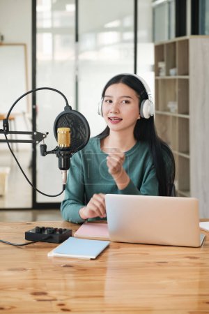 Photo for A woman is sitting at a desk with a microphone and a laptop. She is wearing headphones and she is recording a podcast. The room has a wooden floor and a few books on the desk - Royalty Free Image