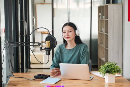 Photo for A woman is sitting at a desk with a microphone and a laptop. She is wearing headphones and smiling. The scene suggests that she is recording a podcast or a voiceover session - Royalty Free Image