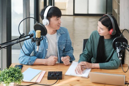 Photo for Two people are sitting at a desk with microphones and a laptop. They are talking to each other and writing something down. Scene is casual and friendly - Royalty Free Image