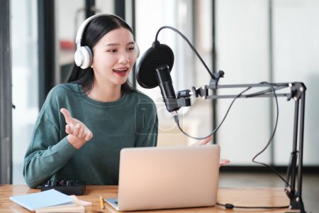 Photo for A woman is sitting at a desk with a laptop and a microphone. She is wearing headphones and she is talking into the microphone. The scene suggests that she is recording a podcast or a video call - Royalty Free Image