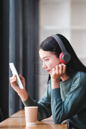 Photo for A woman is sitting at a table with a tablet and a cup of coffee. She is wearing headphones and looking at the tablet - Royalty Free Image