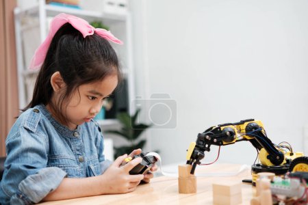 Téléchargez les photos : Une écolière du primaire se concentre sur l'utilisation d'un bras robotisé avec télécommande, démontrant l'éducation aux STIM en action. - en image libre de droit