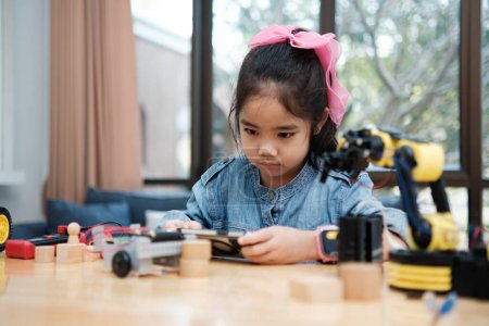 Photo for A young Asian girl in a STEM class attentively uses a smartphone app to remotely control a toy car, showcasing tech education. - Royalty Free Image