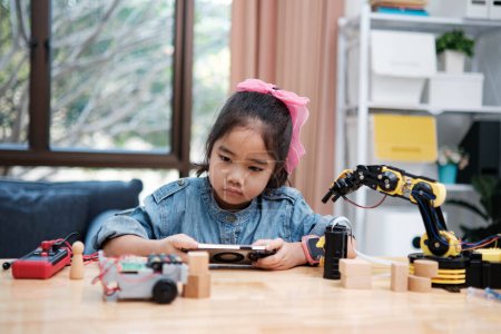 Téléchargez les photos : Une jeune fille asiatique dans une classe de STIM utilise attentivement une application smartphone pour contrôler à distance une voiture jouet, mettant en valeur l'éducation technologique. - en image libre de droit
