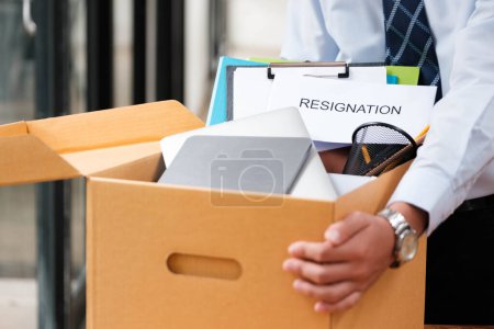 Männlicher Mitarbeiter packt seine persönlichen Sachen in einen Karton mit einem Rücktrittsschreiben, das seine Entscheidung signalisiert, den Job zu verlassen