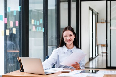 Photo for A cheerful female business professional reviewing documents with a laptop at her workspace in a well-lit office. - Royalty Free Image