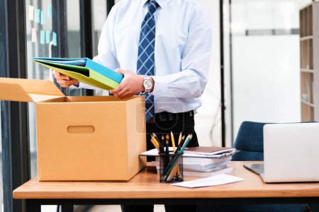 Photo for A man in a suit is opening a cardboard box on a desk. The box contains papers and a laptop - Royalty Free Image