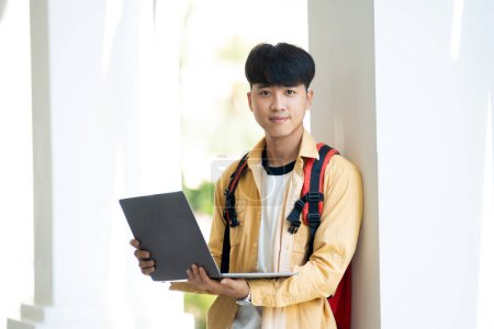 Photo for A male student with a pleasant smile stands in the school hallway, confidently holding his laptop, ready for class. - Royalty Free Image