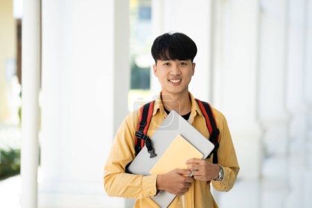 Photo for A college student stands in the hallway of his school, holding textbooks and smiling, ready for a day of learning and studies. - Royalty Free Image