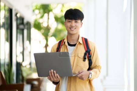 Téléchargez les photos : Un étudiant homme avec un sourire agréable se tient dans le couloir de l'école, tenant avec confiance son ordinateur portable, prêt pour la classe. - en image libre de droit