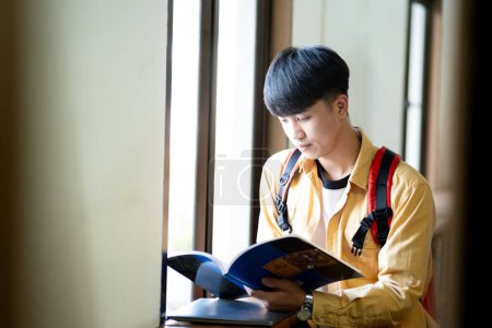 Photo for A young man is reading a book while wearing a yellow shirt and a red backpack. He is sitting by a window, and the book he is reading is blue - Royalty Free Image