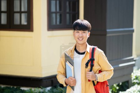 Photo for A happy and content young man stands outside on his university campus, holding a laptop and books, embodying the optimism of college life. - Royalty Free Image