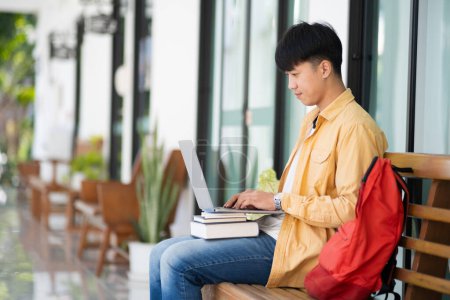 Téléchargez les photos : Un étudiant est assis sur un banc extérieur, travaillant intensément sur son ordinateur portable, avec une pile de livres et un sac à dos rouge à ses côtés. - en image libre de droit