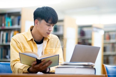 Photo for A young man is sitting at a table in a library, reading a book and using a laptop. Concept of focus and concentration, as the man is engaged in his studies. The library setting suggests a quiet - Royalty Free Image
