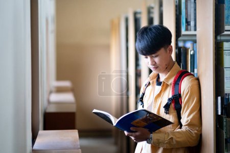 Photo for A young man is reading a book in a library. He is wearing a backpack and has a serious expression on his face. The library is quiet and peaceful - Royalty Free Image