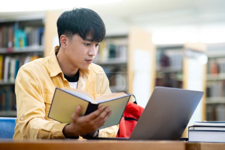 Photo for A young man is sitting at a desk with a laptop and a book. He is reading the book while looking at the laptop. The scene suggests that he is studying or working on a project - Royalty Free Image