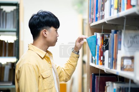 Photo for A man in a yellow shirt is looking at a book on a library shelf. He is reaching for a book with a blue cover - Royalty Free Image