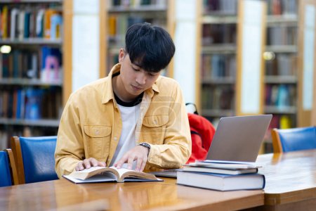Photo for A young man is sitting at a table in a library, reading a book. He is wearing a yellow shirt and a black shirt. There are several books on the table, including a laptop - Royalty Free Image