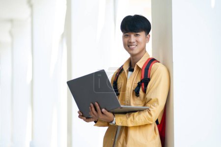 Foto de Un estudiante masculino con una sonrisa agradable está parado en el pasillo de la escuela, sosteniendo con confianza su computadora portátil, listo para la clase. - Imagen libre de derechos