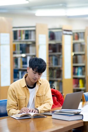 Photo for A young man is sitting at a table in a library, reading a book and using a laptop. Concept of focus and concentration, as the man is engaged in his studies. The library setting suggests a quiet - Royalty Free Image