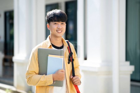 Photo for A contented college student carrying a laptop and books walks across the campus grounds, exuding a sense of readiness and enthusiasm for learning. - Royalty Free Image