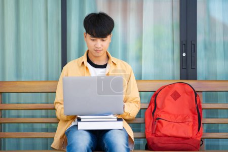 Photo for A college student sits on an outdoor bench, working intently on his laptop, with a stack of books and a red backpack by his side. - Royalty Free Image