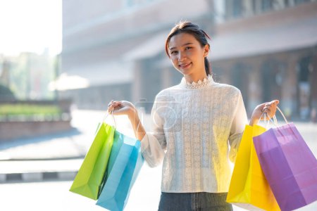Téléchargez les photos : Joyeux jeune femme profitant d'une virée shopping, tenant des sacs à provisions colorés dans un cadre extérieur ensoleillé. - en image libre de droit