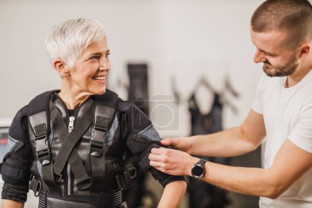 Foto de Entrenador personal está ayudando a una mujer de mediana edad con traje de vestir para EMS hacer ejercicio en el gimnasio. - Imagen libre de derechos