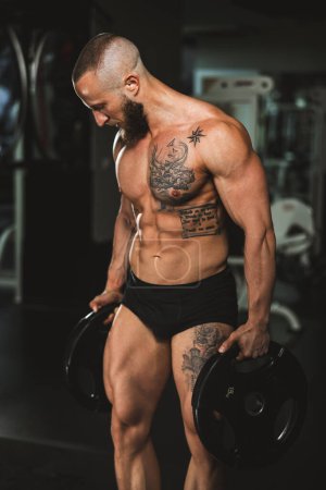 Téléchargez les photos : Prise de vue d'un bodybuilder musculaire montrant ses muscles parfaits après avoir travaillé avec la plaque de poids à la salle de gym. - en image libre de droit