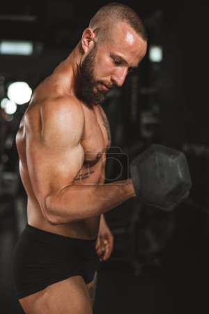 Téléchargez les photos : Shot of a muscular bodybuilder doing hard training with dumbbell at the gym. He is pumping up his biceps muscle with heavy weight. - en image libre de droit