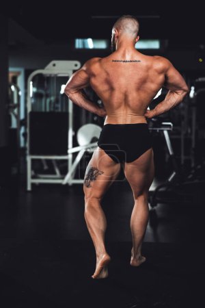 Téléchargez les photos : Vue arrière d'un jeune bodybuilder musculaire montrant ses muscles parfaits du dos tout en posant après avoir travaillé à la salle de gym. - en image libre de droit