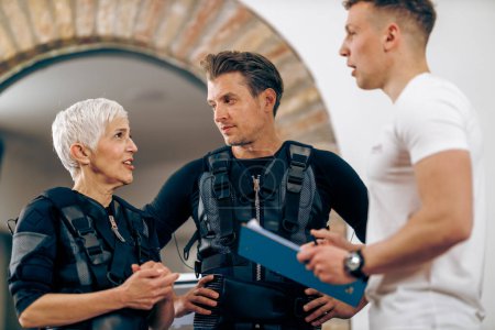Foto de A senior mother and her middle age son talking with personal trainer during EMS workout in the gym. - Imagen libre de derechos