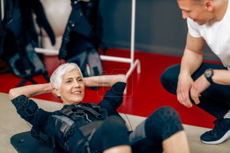 Foto de Senior woman is doing sit-up exercises during EMS training with personal trainer in the gym. - Imagen libre de derechos