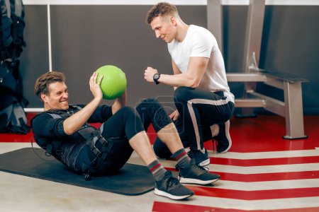 Photo for Middle age man is doing sit-up exercises during EMS training with personal trainer in the gym. - Royalty Free Image