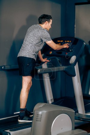 Téléchargez les photos : Tourné d'un homme d'âge moyen se prépare à courir sur un tapis roulant à la salle de gym. - en image libre de droit
