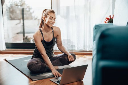 Téléchargez les photos : Jeune femme utilisant un ordinateur portable tout en faisant de la formation de conditionnement physique à la maison. - en image libre de droit