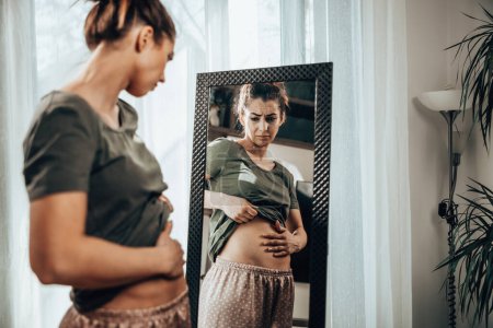 Photo for Frown young woman standing in front of a mirror and holding hands on her bloating stomach. - Royalty Free Image